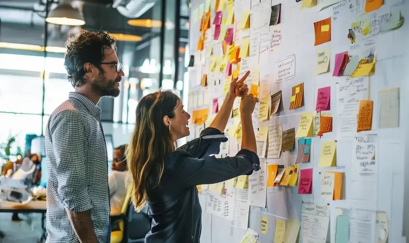 Two coworkers stand closely together reviewing a large whiteboard filled with colorful sticky notes, sketches, and handwritten plans in a modern, industrial-style office.