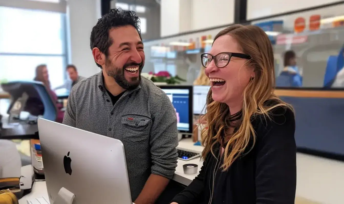 Two coworkers are sharing a laugh while working at a computer in a bright, open office space. The man and woman are clearly enjoying the moment, with genuine smiles and relaxed energy, suggesting a fun and collaborative workplace culture.