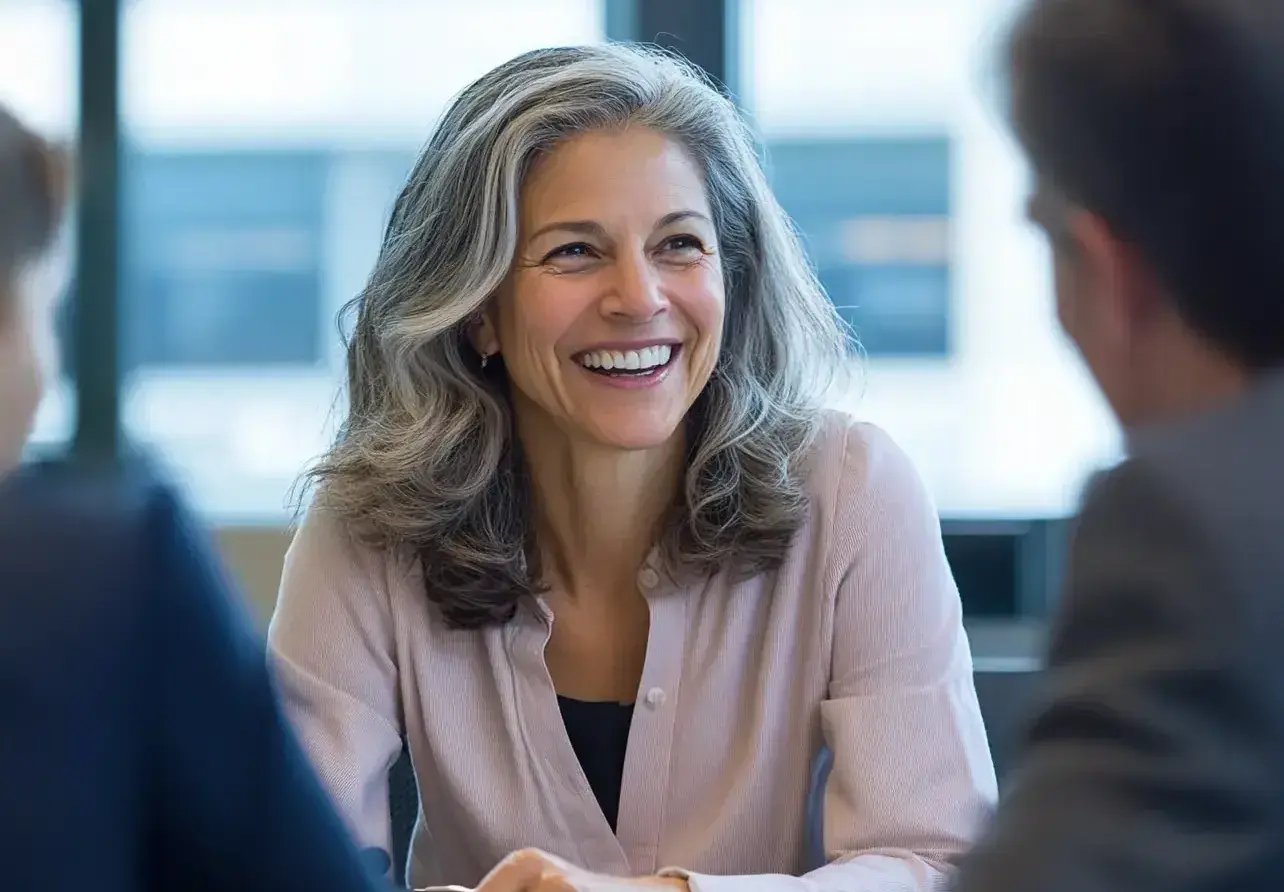 A smiling woman with long, wavy gray hair and a light pink blouse is engaged in a conversation with two colleagues across a table in a bright, modern office. Her confident, open expression reflects a positive and collaborative work environment.