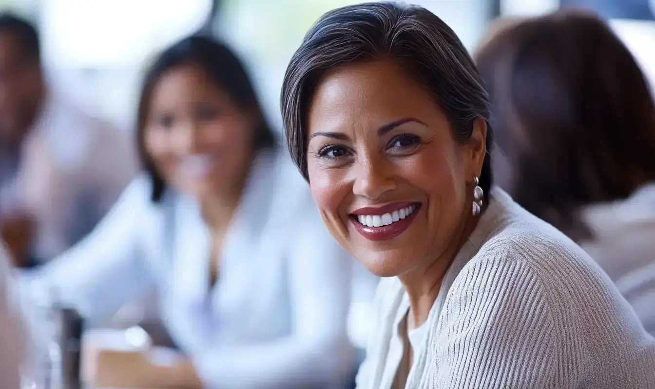 A confident woman with short dark hair and pearl earrings is smiling warmly while seated at a table during a meeting or group discussion. The blurred background shows others engaged in conversation, suggesting a positive and collaborative environment.