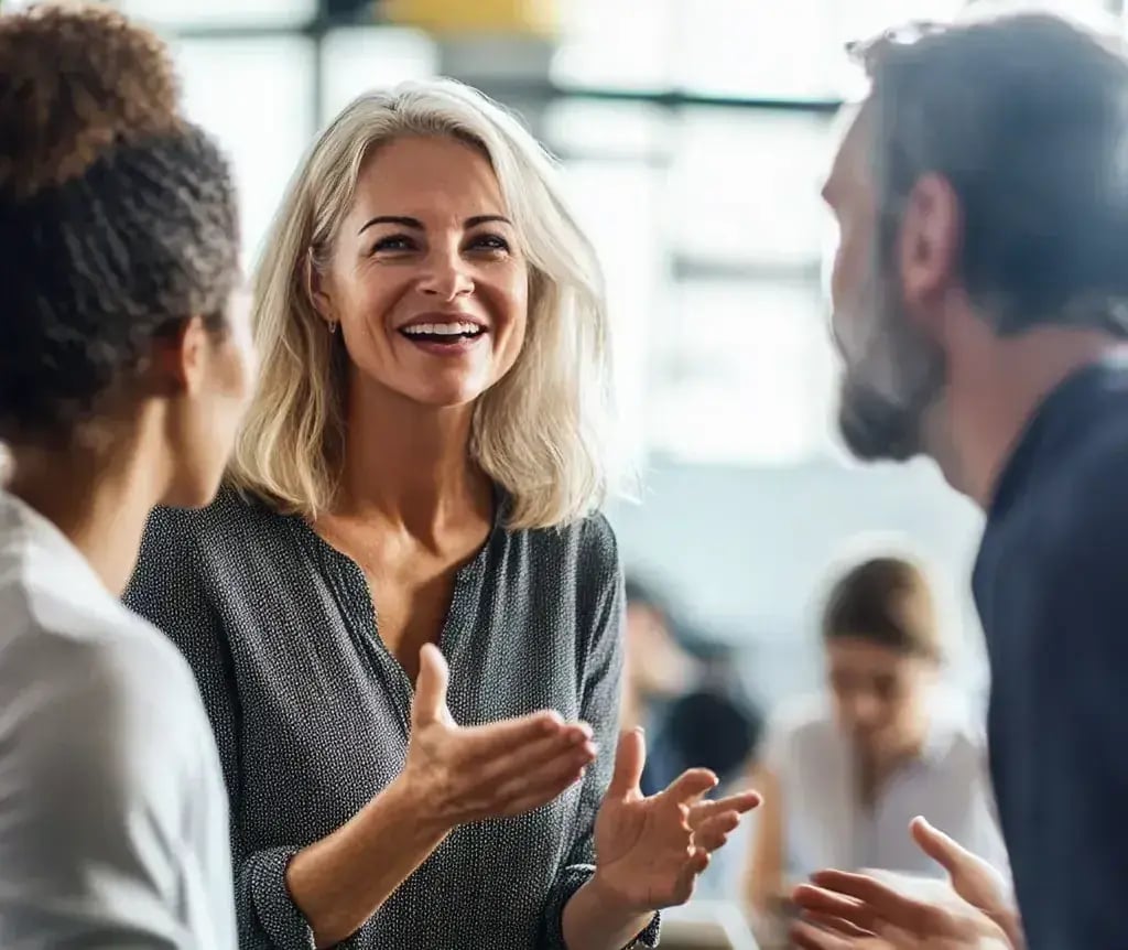 A professional woman with shoulder-length gray-blonde hair is smiling and gesturing as she chats with two coworkers in a modern office setting. The atmosphere is casual and friendly, with blurred background figures suggesting an open, collaborative workspace.